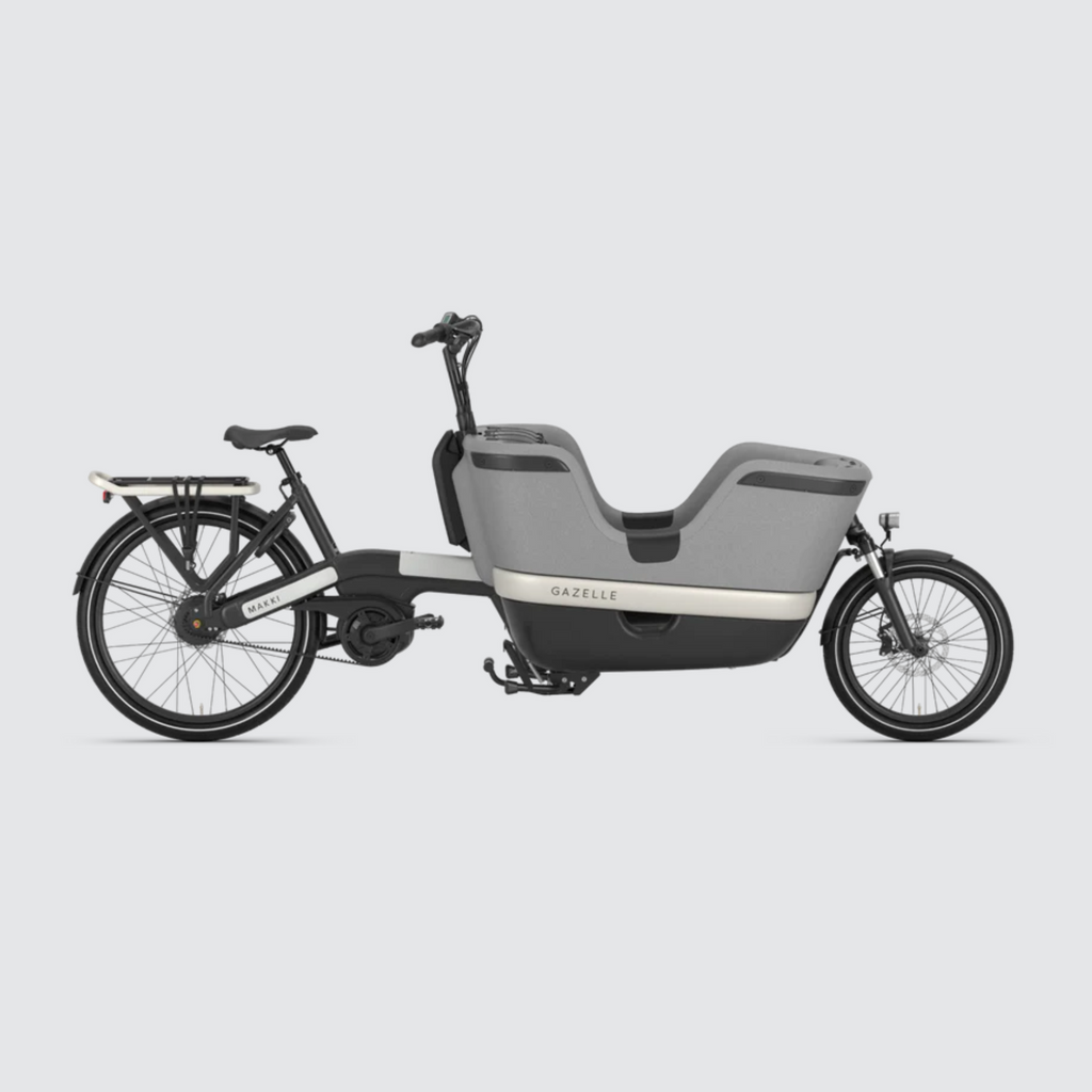 A side view image of the Gazelle Makki HMB cargo ebike, showing the full length of the bike. At the rear is a pannier rack with bungee strap, and a belt drive providing less maintenance and quieter riding. The high EPP box is suitable for carrying two children with safety belts or large loads. With a reflector fixed to the rear rack system and an integrated LED lamp on the front, the Makki Load Cargo includes an integrated battery pack and a double kicstand for loading stability. 